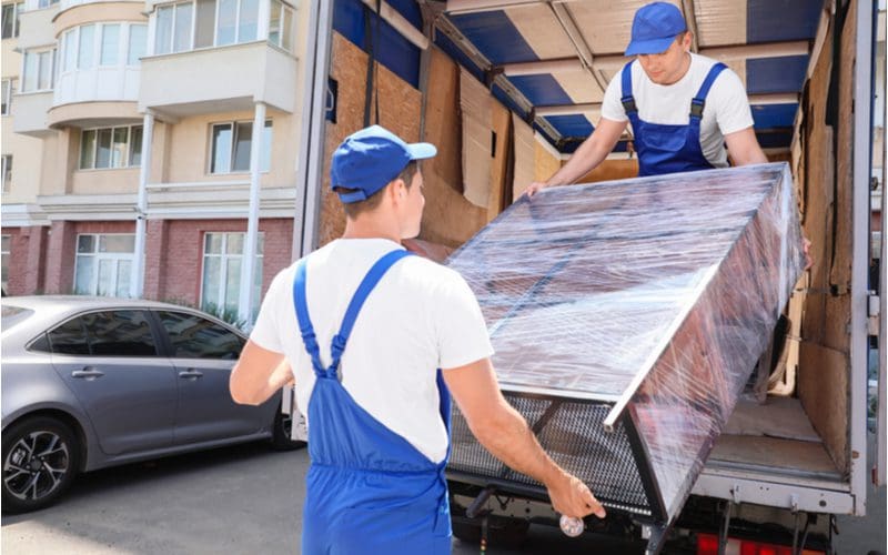 Two men in white shirts with blue overalls and hats pulling a well-wrapped black metal shelf from a truck for a piece on whether or not you should tip furniture delivery men