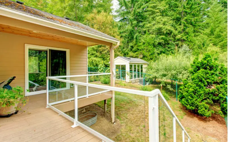 Walkout deck with glass railings with white frames in the back of a heavily forested lawn