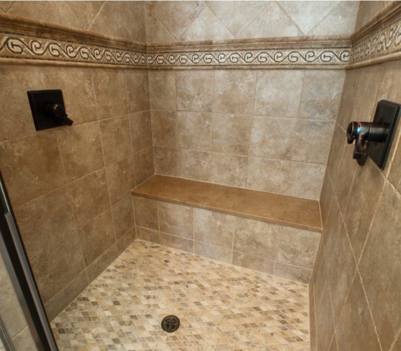 Tile shower idea featuring a big square shower with a bench surrounded by travertine tile in a dark tan color