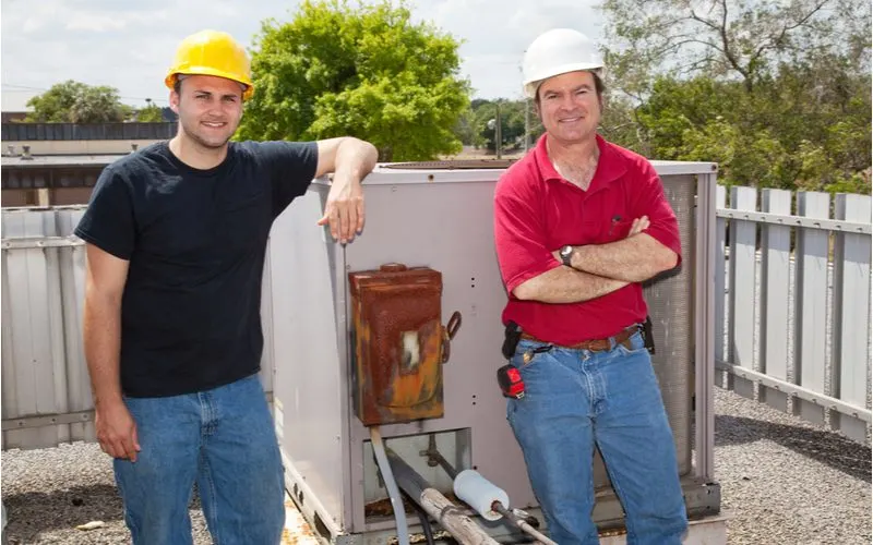 Two ac repair men bidding out the cost of a new hvac installation while standing in front of an old unit