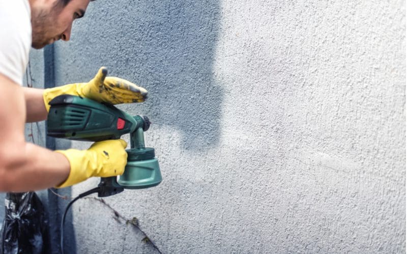 Guy painting the exterior of a home with a spray gun and grey paint
