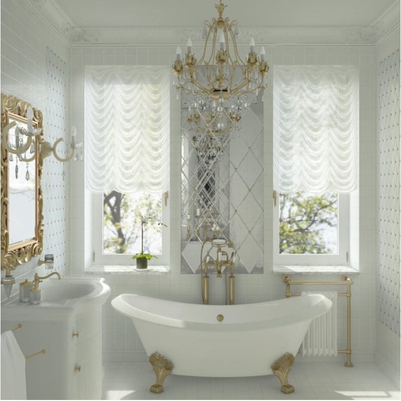 White clawfoot tub in a white room with two large windows on either side of the tub below a glass and gold chandelier