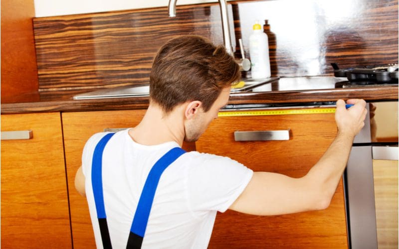 Guy using a tape measure to determine the average dishwasher size in preparation to install a new one in a nice modern woody cabineted kitchen
