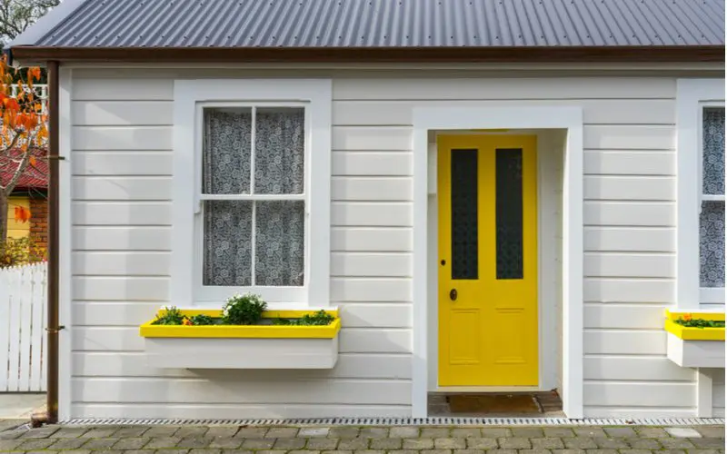 For a piece on the best front door colors for tan houses, a tan house with a bright yellow door with yellow-trimmed flower boxes and a metal roof