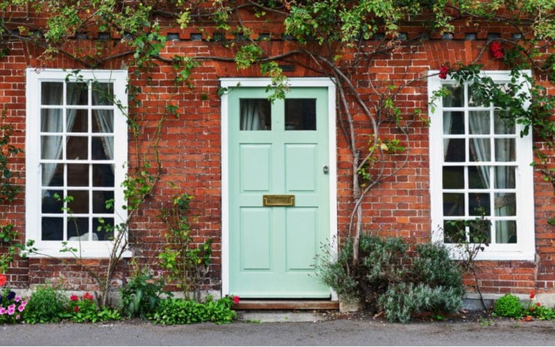 Eggshell green, one of the most unique front door colors for red brick houses, on a colonial-style home with plants in the front