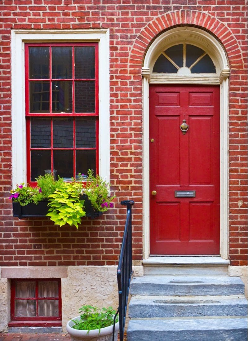 Bright red door on a red brick house next to a painted cement staircase and black railing with a window planter to the left