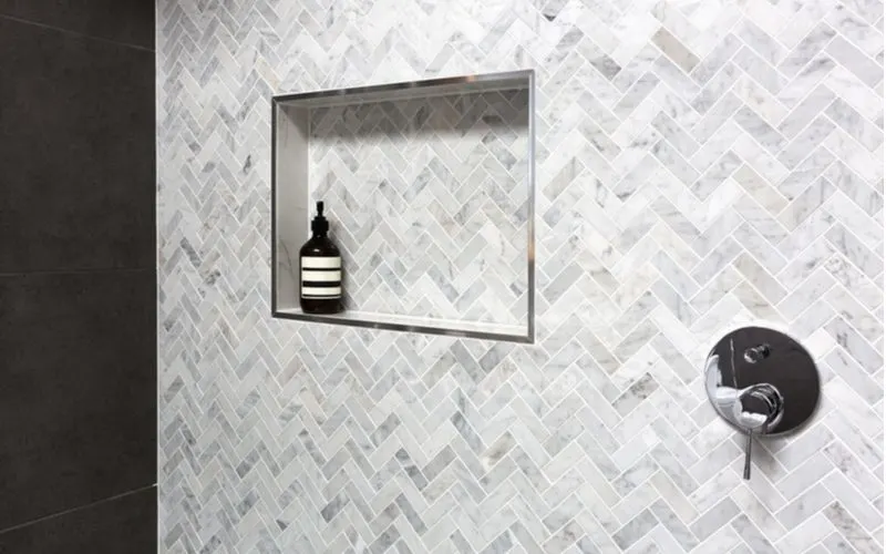 Grey tile bathroom shower surround idea with herringbone tile with chrome fixtures and a chrome trimmed soap shelf