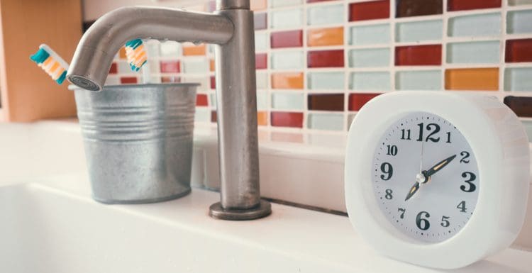 Featured image for a piece titled Clock in Bathroom featuring a small analog clock on a vanity next to a single handle faucet
