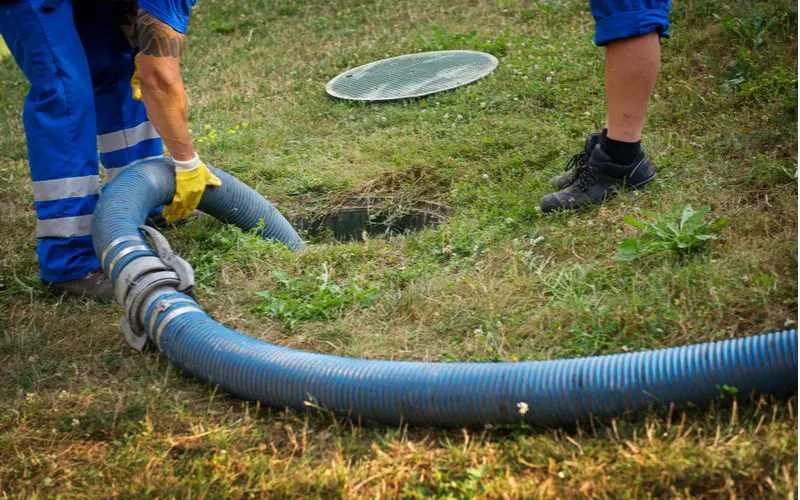 Man emptying a septic tank with a big hose while another guy in black shoes stands over and watches