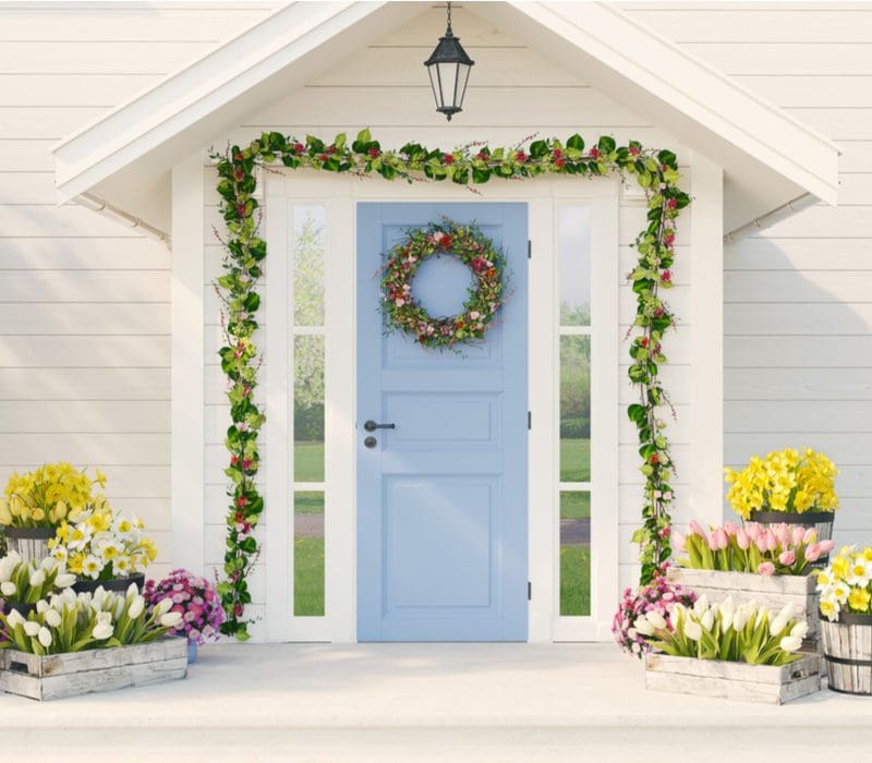 Carolina blue front door color on a tan house with garland wrapped around the top and Spring flowers and tulips in boxes and barrels on the sides of the porch