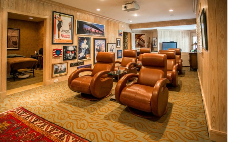 Man cave idea featuring swirly gold and beige carpet, a number of brown leather reclining theatre seats, and vertical natural wood-paneled walls
