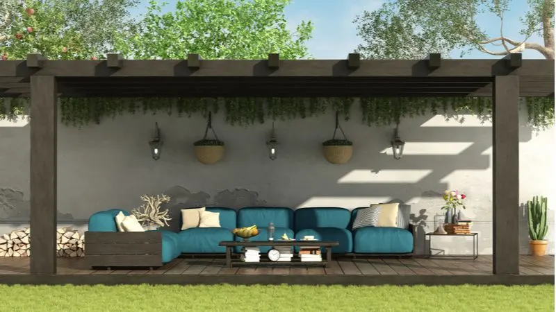 Large beam pergola idea with dark wood and thick lumber attached to a stucco wall below which sits a slightly elevated wooden deck with no railings and logs and teal patio furniture
