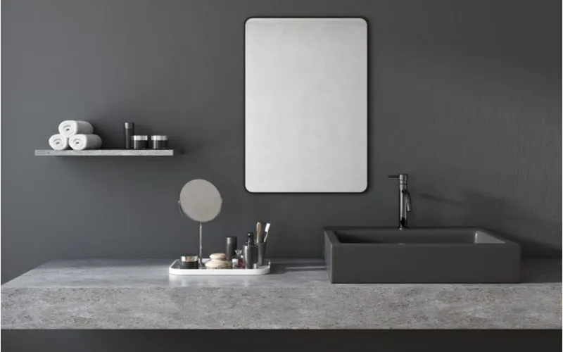 Bathroom shelf idea with a stone-look floating shelf mounted to a dark grey wall next to a mirror and above the stone vanity top
