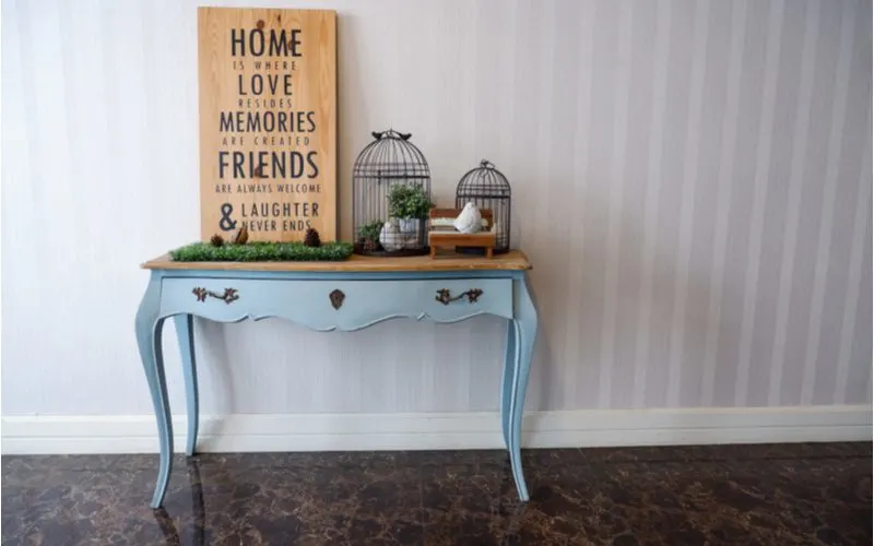 Farmhouse style entryway table decor idea with a mint green table with a natural wood top holding up a rustic and corny wooden sign and two birdcages with some natural greenery accents