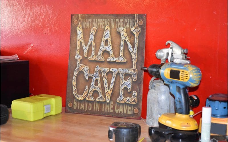 Image of a man cave sign leaning against a red wall next to power tools on a rustic-looking shelf