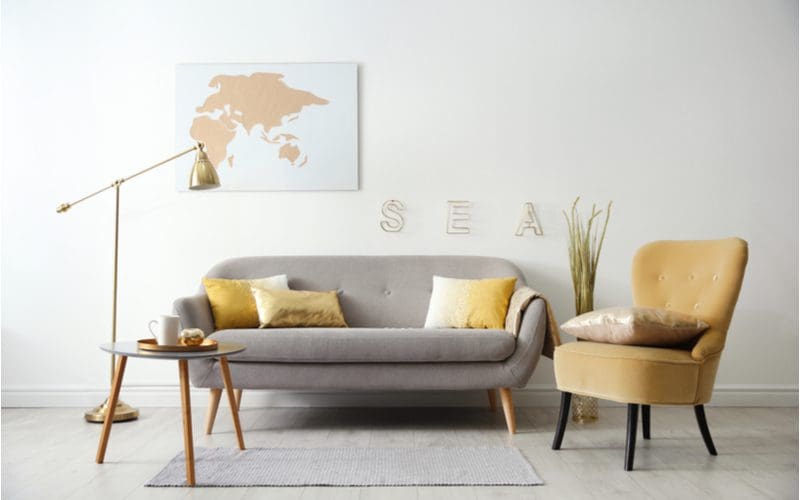Wall decor above couch idea featuring a white wall with a grey couch and very light brown accent pillows and chair with a world map in printed gold paint above the couch