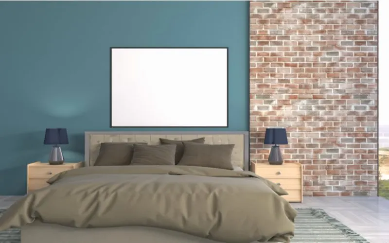 A blank framed canvas hangs on the wall above a brown bed to the left of a brick wall for an idea for a men's bedroom