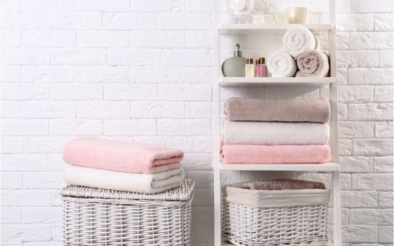 For a piece on bathroom towel storage ideas, a white rack sits next to a white wicker basket on which towels sit in front of a white brick wall