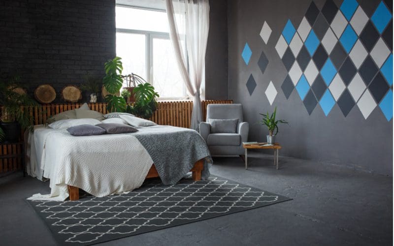 Mens bedroom idea showing an otherwise dark grey room with white and blue square tiles stuck to the wall and a bed with a white blanket over a dark grey patterned rug