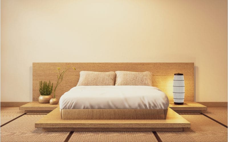 Photo of a light wood bed frame and bed base sitting on a raised platform with a Japanese-style plant to the left and an oval light to the right