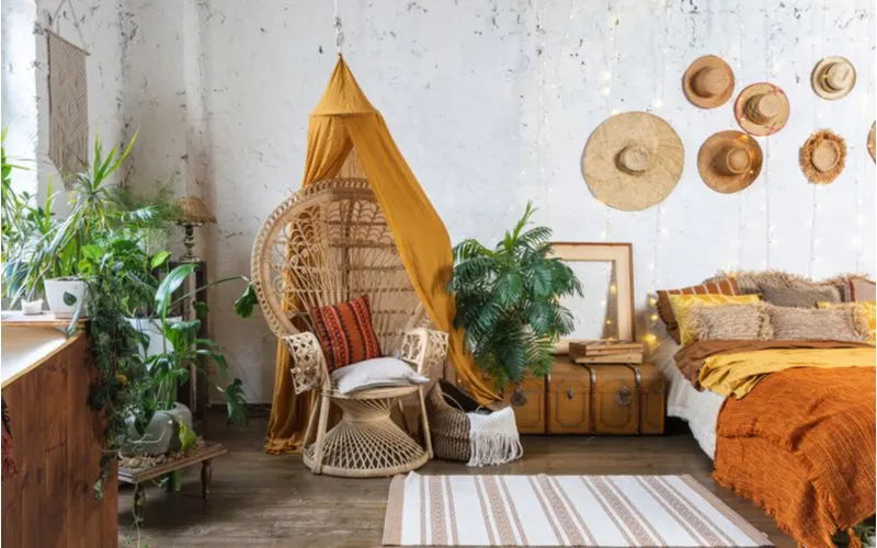 Image for a piece on boho bedroom ideas featuring a cozy interior with a wicker chair, green plants, and lots of wicker hats and furniture on the walls and floor with a trunk as a side table
