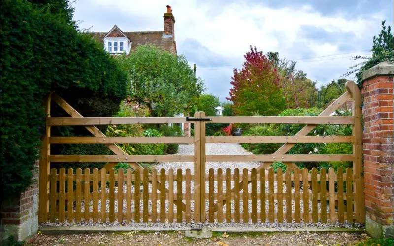 Farm style wooden driveway gate idea with both vertical and horizontal natural wooden slats between two pillars made of red brick with light grey concrete toppers