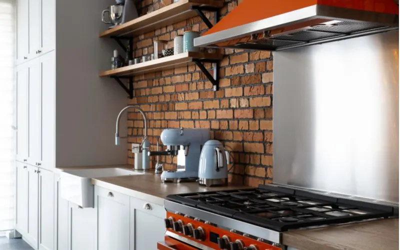 Rustic red brick textured farmhouse backsplash lining the wall of a simple home with stainless and red appliances with a farmhouse sink and very faint teal cabinets