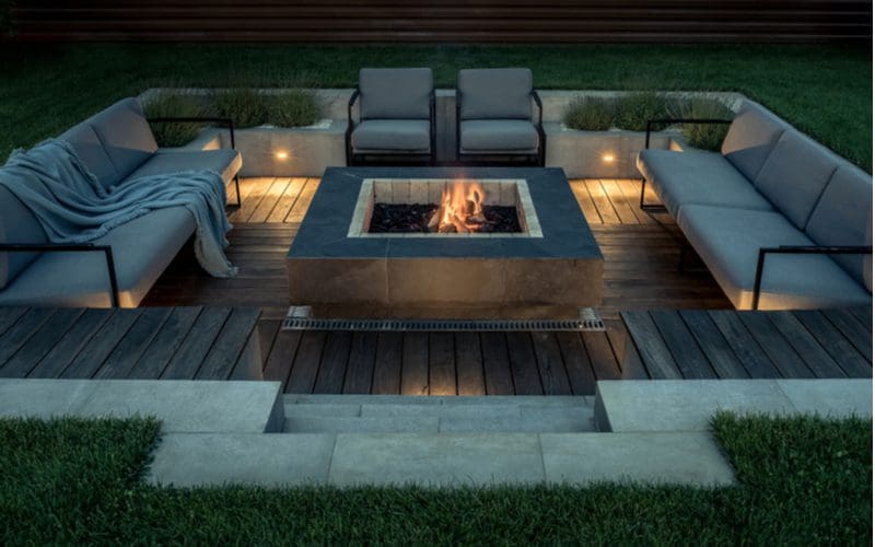 Fire pit idea with a sunken square concrete paver fire pit with wood decking supporting metal padded chairs