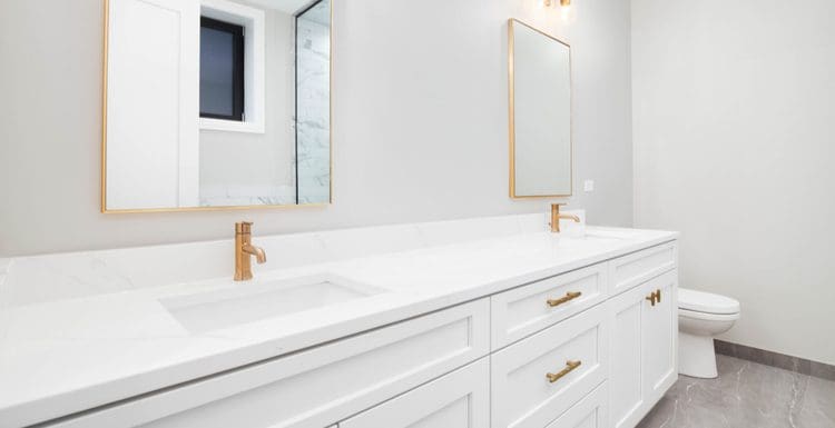 Image of a white and gold bathroom idea for a piece on this topic with white walls, toilet, vanity, vanity top, grey tile, and gold fixtures, mirrors, lights, and hardware
