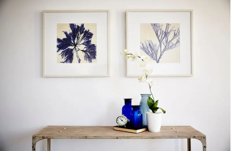 Entry table decor idea titled color coordinate your art with a white entryway wall from which hang two beige framed plant prints above an entry table in reclaimed wood with blue and teal decorations