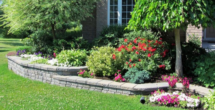 Flower bed ideas featuring a number of paver blocks that line a shrub and tree and flower bed outside a brick house