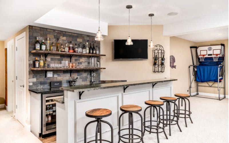 Man cave idea titled "fully stocked bar" featuring a wet bar with wooden topped stools and a basketball shooting machine right by a TV mounted on the wall