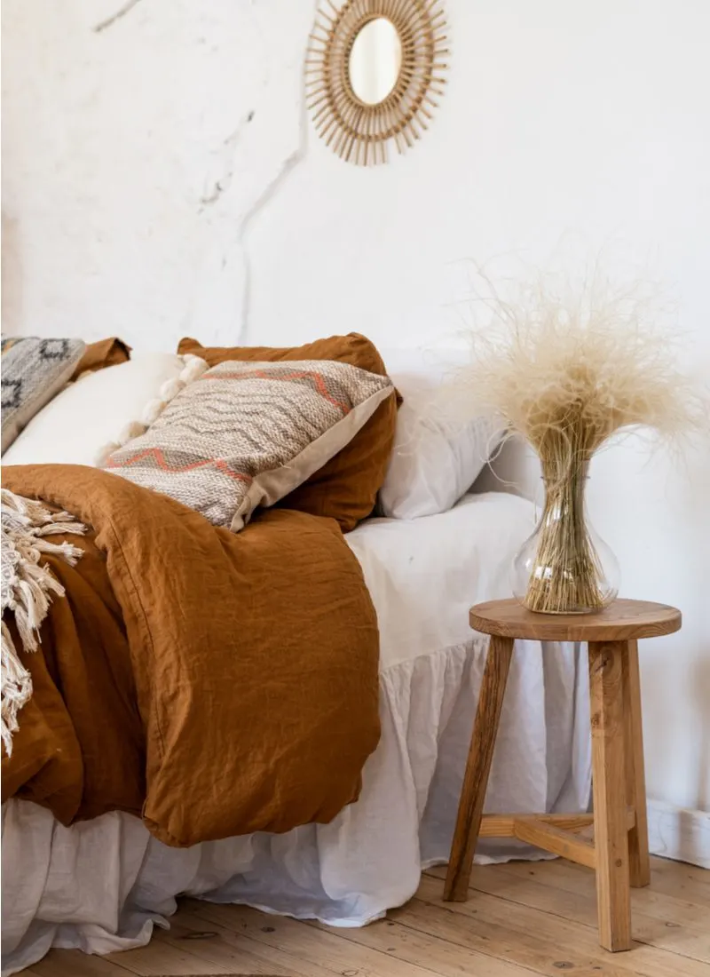 Bohemian style bedroom with dark orange-brown style bedspread next to a natural wooden table below a round metal sun-type art fixture with a mirror in the middle hanging on the wall