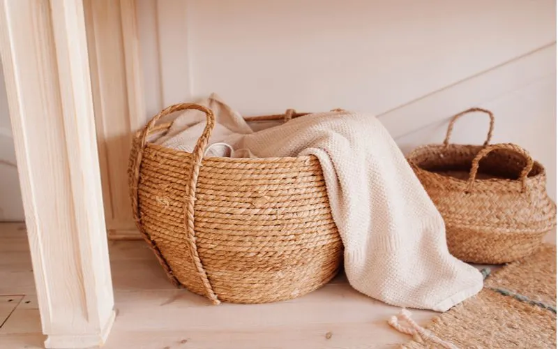 For an idea for boho bedroom decorations, a weaved basket in light brown coloring sits on a light grey wooden plank floor holding a white blanket that looks very cozy