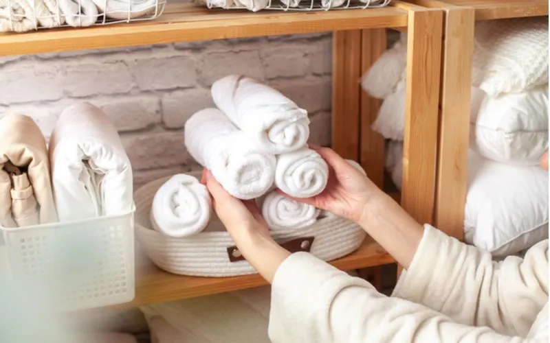 For an idea for small bathroom storage, a woman rolls her towels and places them in an open wooden shelving unit