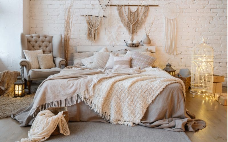 Boho bedroom featuring a fluffy comforter and blanket draped over the bed with mostly grey and brown furniture and accents with lots of tapestries hanging on the wall next to a bird cage around which are string lights