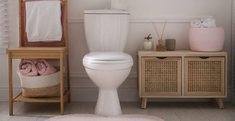 For a piece titled Standard Toilet Dimensions, a standard height elongated toilet sits on a grey wood plank-looking tile floor between a wooden chair and a towel storage chest