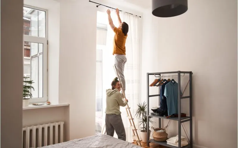 For a frequently asked section for a piece on should curtains touch the floor, a man hangs curtains while his partner supports the ladder