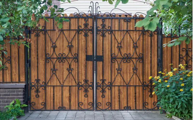 Ornate hybrid metal gate with light natural wood slats in back above a paver driveway between two trees