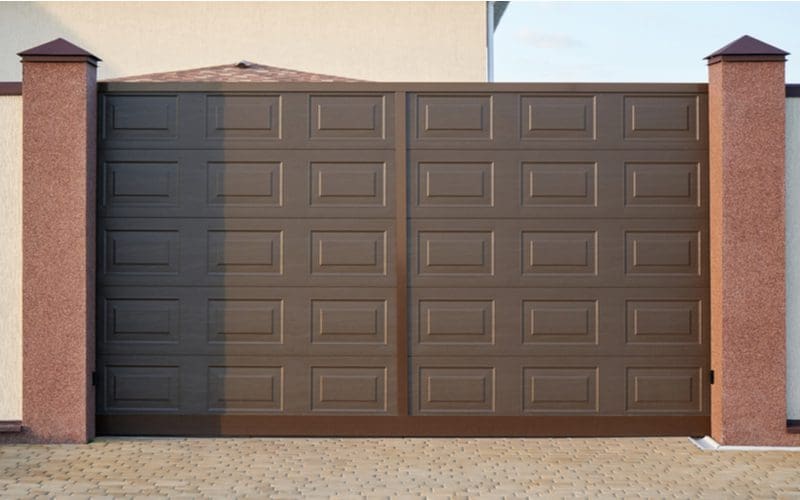 Image for a roundup on driveway gate ideas featuring a driveway gate made out of brown pvc between two concrete or stone pillars skinny in size