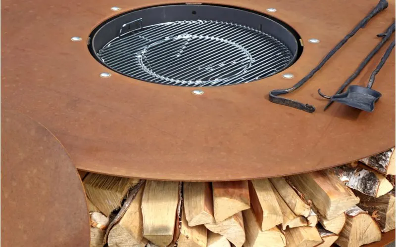 A wooden fire pit idea with wood storage shelves below the metal firepit on which sits a poker and ash shovel