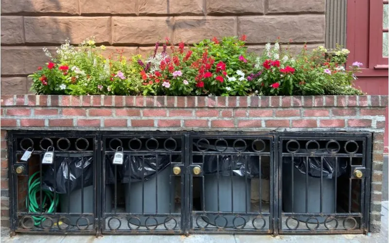 Urban-style front yard landscaping idea in which trash cans are hidden behind a brick wall with built-in planter behind a locked gate