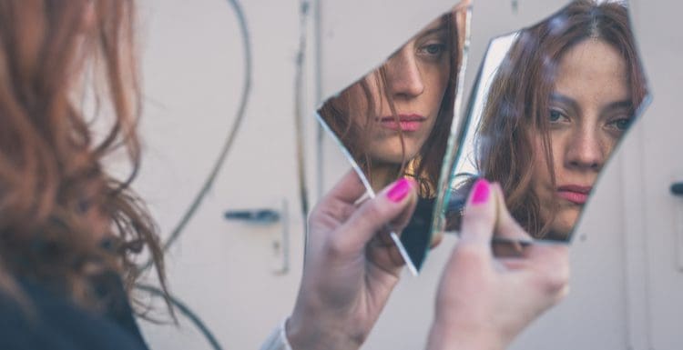 Featured image for a piece on how to fix a broken mirror featuring a woman looking into two pieces of a broken mirror and seeing her reflection