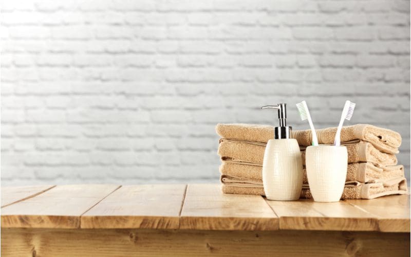 Close-up shot of a bathroom shelf made of natural wood on which sit toothbrushes, towels, and soap in front of a white painted brick wall