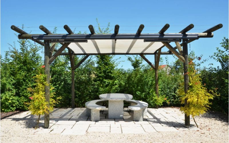 Black log pergola idea with a beige sunshade suspended over a round stone picnic table sitting on a cement tile patio