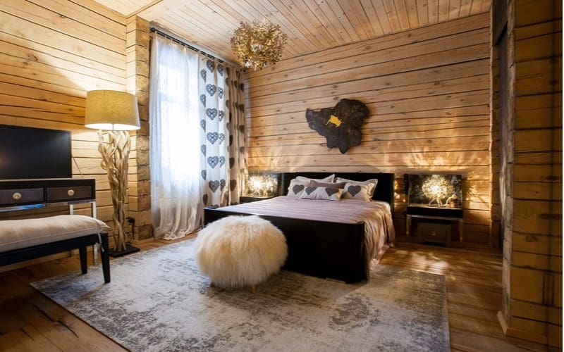 Country aesthetic bedroom idea with natural wood paneling on the walls and ceiling with large barnwood flooring panels and ample natural wood lamps, chandeliers, and décor alongside animal hides and fur on the wall and furniture