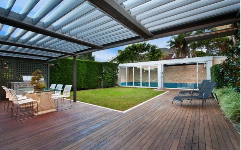 Pergola idea showing a partially covering a wooden slatted patio with a walkway to the small poolhouse in back