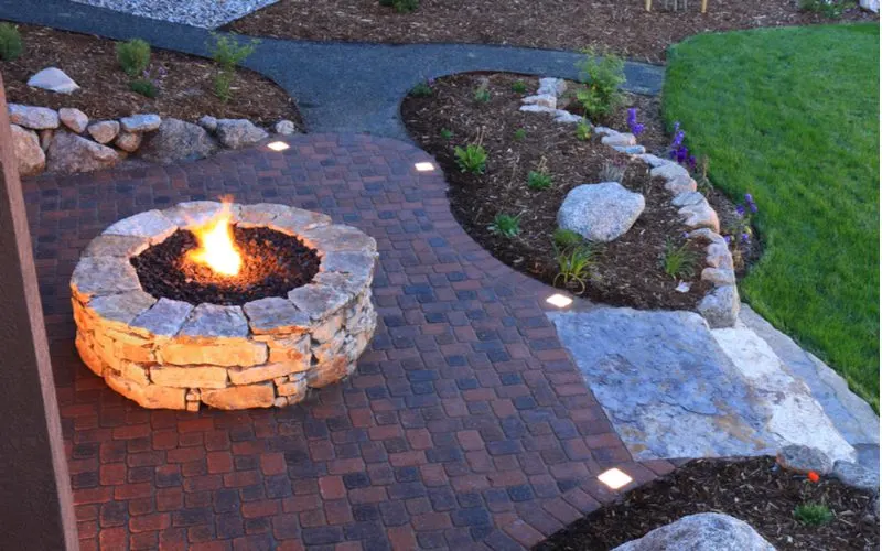 Gas fire pit idea with lava rock built out of flagstone block in the middle of a dark square paver patio