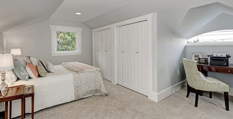 Featured image for a roundup on unique closet door ideas featuring a dormer bedroom with double white bi-fold closet doors next to a small work station and a bed