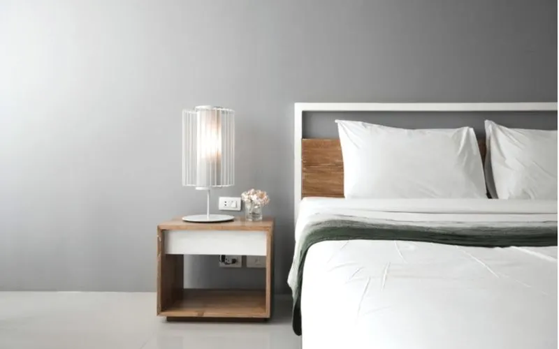 A no-nonsense bedside table with an open layout and white accent trim piece sits to the left of a white open-frame bed in a grey men's bedroom